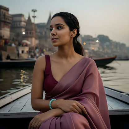Woman in a pink saree sitting by a river with a cityscape in the background